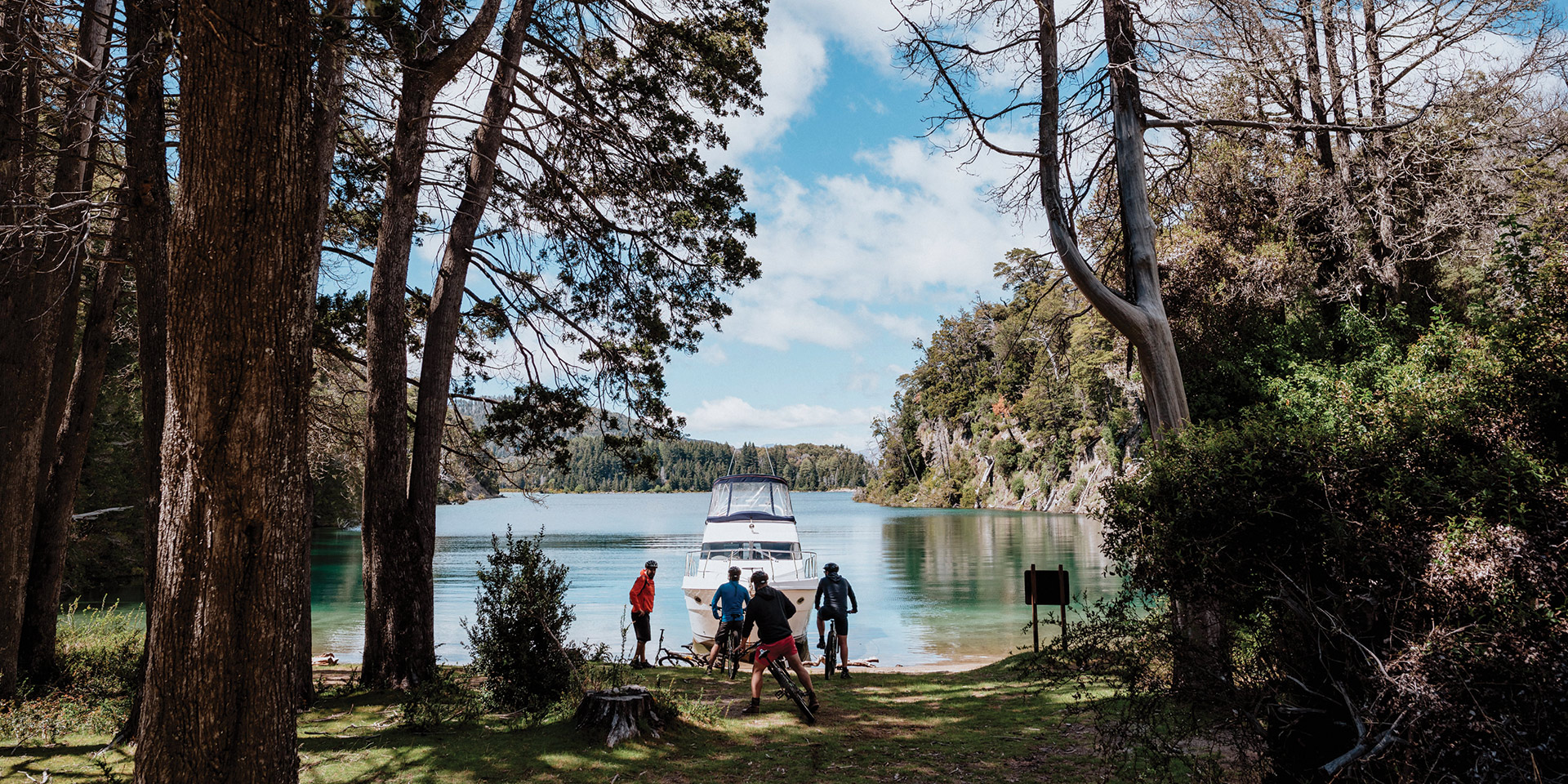 A scenic view of a group of people with bicycles and a boat on the shore of a calm lake in Patagonia's Lake District. Tall trees frame the foreground, and forested hills rise from the opposite shore under a partly cloudy sky.