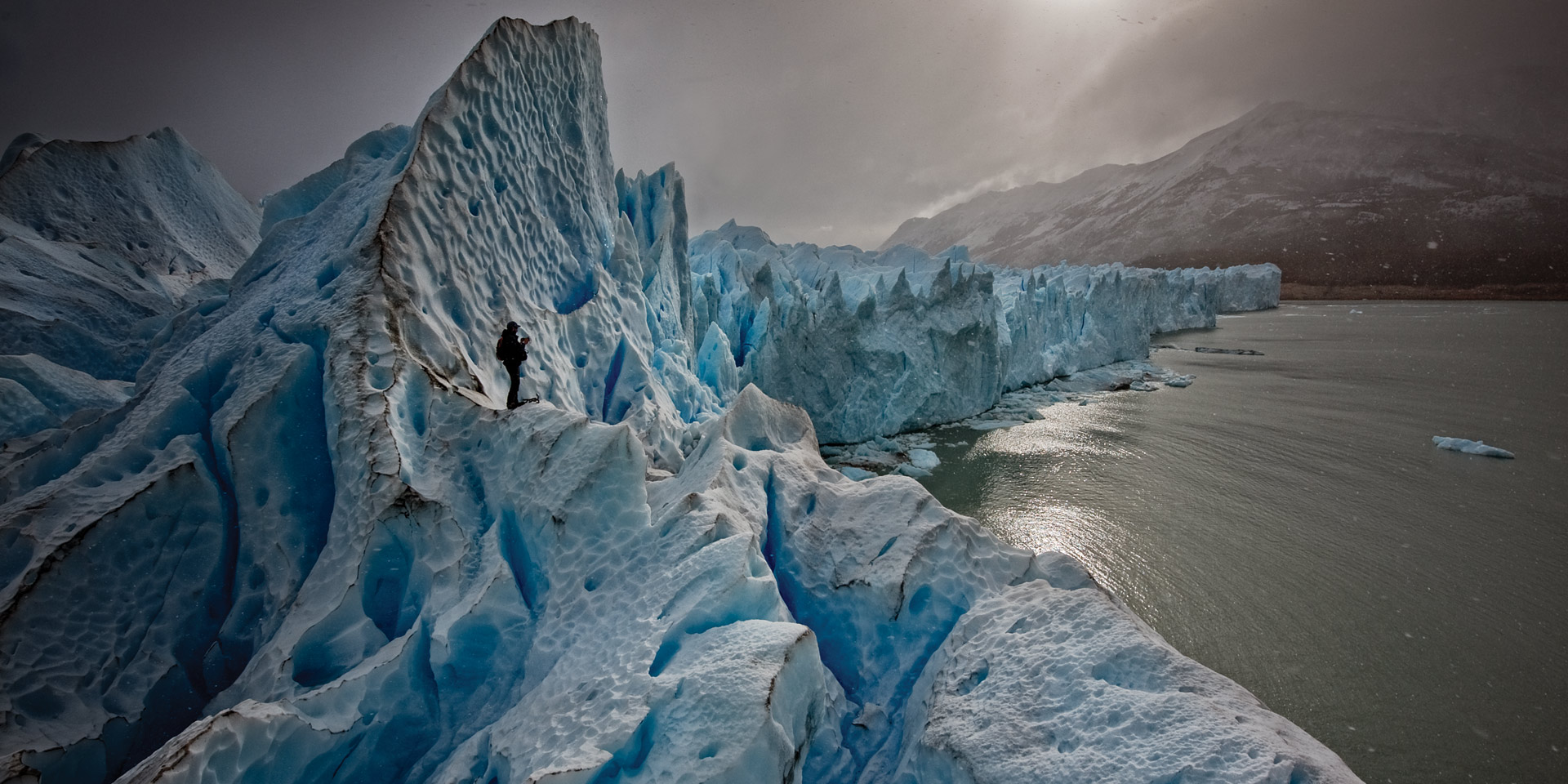 A dramatic view of a vast, jagged glacier in Los Glaciares National Park, with a small figure of a person providing scale. The glacier meets a grey lake under a cloudy sky, with distant mountains visible