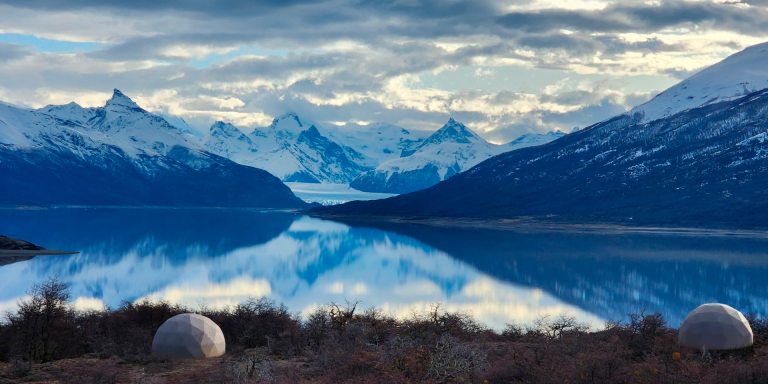 Two white, geodesic domes nestled among vegetation, with a stunning view of Perito Moreno Glacier, a glacial lake and snow-capped mountains.