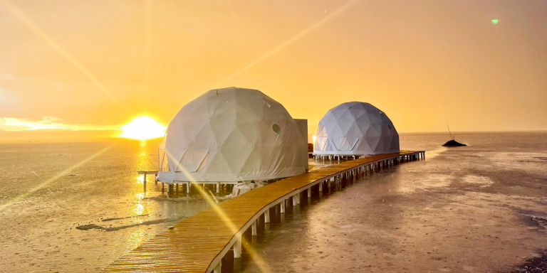 Two geodesic domes on a wooden walkway extending into a shallow body of water, with a vibrant sunset in the background.
