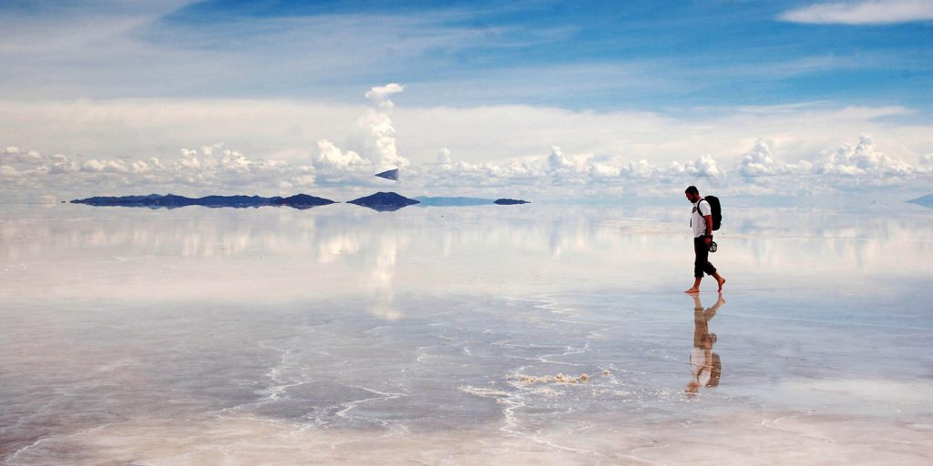 A lone traveler walks across the vast, reflective surface of a South American salt flat under a blue and cloudy sky. Distant landmasses are visible on the horizon, mirrored in the salt.