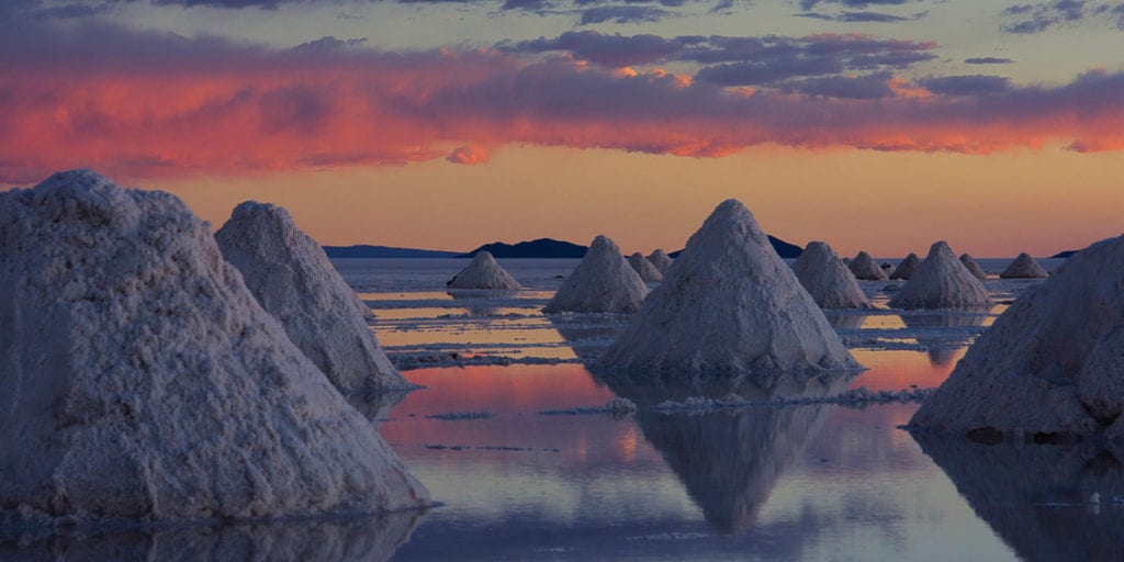 Salt mounds reflected in the water on the Uyuni Salt Flats at sunset, with a colorful sky in the background.