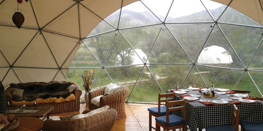 The interior of a geodesic dome with a dining table, chairs, and sofas, overlooking a scenic landscape.