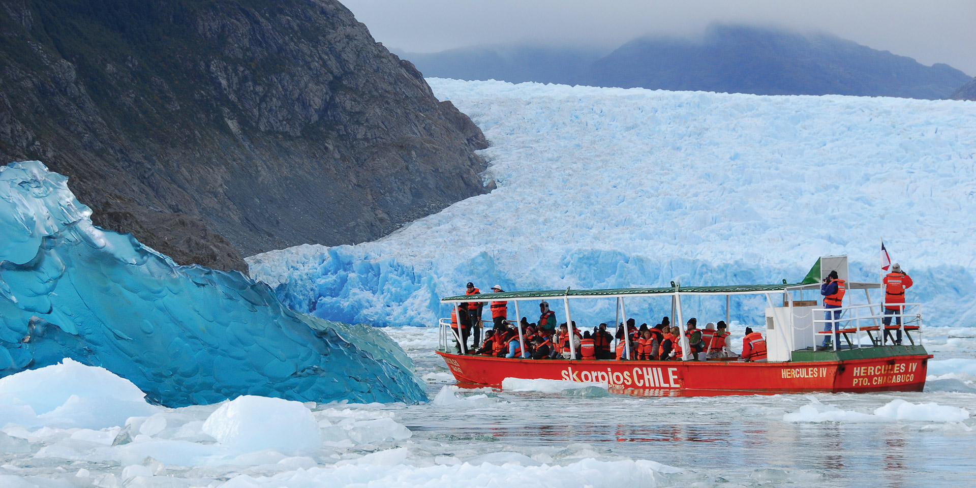 An image of a red aboat in a fjord filled with ice floes, with a large glacier and rugged mountains forming a dramatic backdrop.
