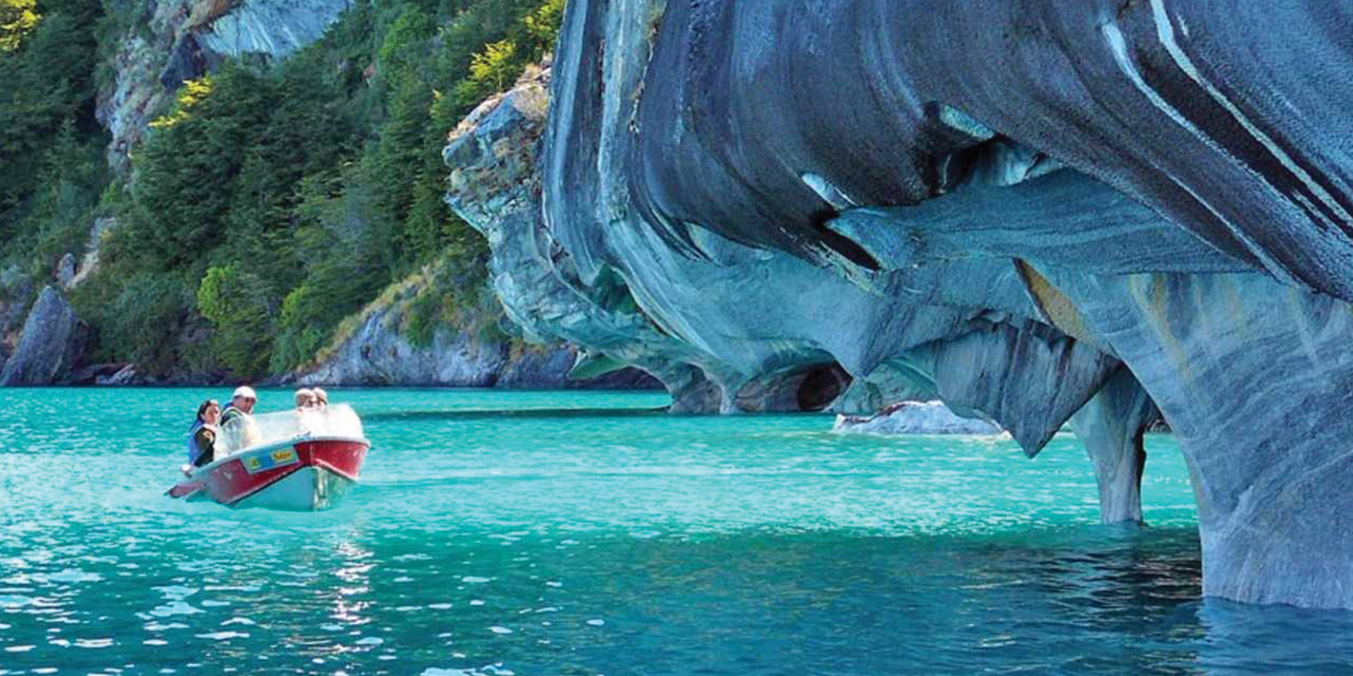 A stunning view of the Marble Caves, unique geological formations with swirling blue and grey patterns, reflected in the turquoise waters of General Carrera Lake. A small boat with tourists is visible.