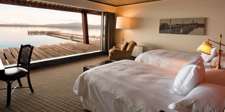 Interior view of a room at Singular Patagonia, featuring two beds, a chair, and a large window overlooking a lake and dock.