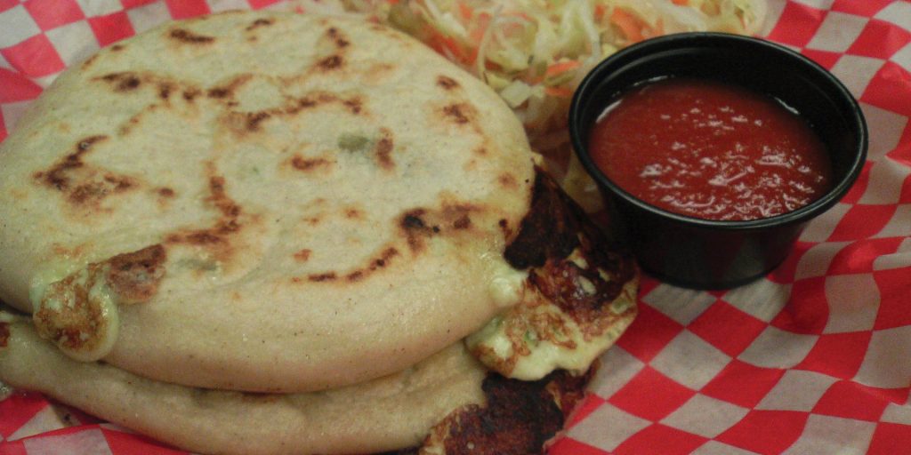 Two golden-brown arepas filled with cheese and other ingredients, served with curtido and salsa on a red and white checkered paper.