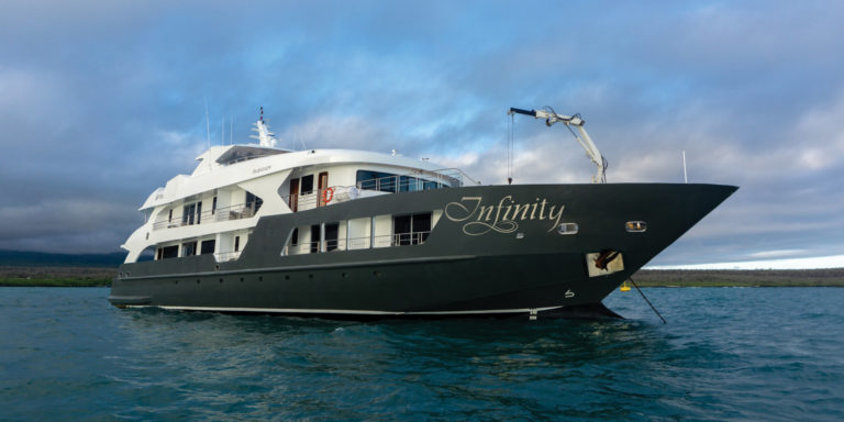 Exterior view of the Infinity Yacht in the Galapagos Islands, showing the dark grey hull, white upper deck, and "Infinity" logo.