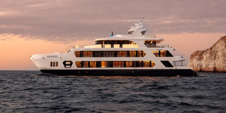 The image showcases the Hermes Mega Catamaran, a sleek and modern vessel with a predominantly white hull and a navy blue base. It's positioned in open water, likely at sunset, with a warm glow illuminating the sky. The catamaran's design emphasises spaciousness and comfort, with multiple decks and large windows.
