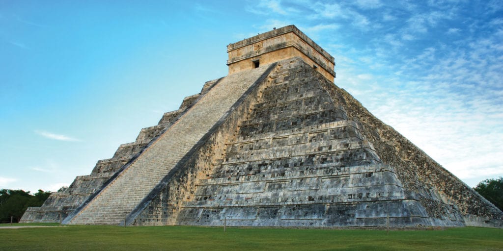 A large, stepped pyramid made of gray stone stands in a grassy field under a blue sky with wispy clouds.