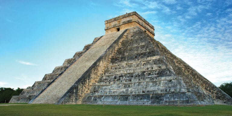 A large, stepped pyramid made of gray stone stands in a grassy field under a blue sky with wispy clouds.