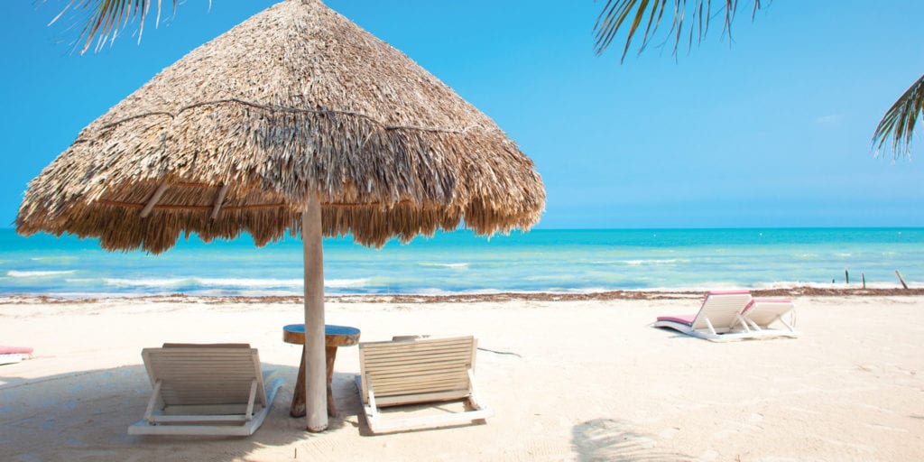 A white sandy beach with several lounge chairs and thatched umbrellas, overlooking calm turquoise water under a clear blue sky with palm tree fronds visible in the corners.