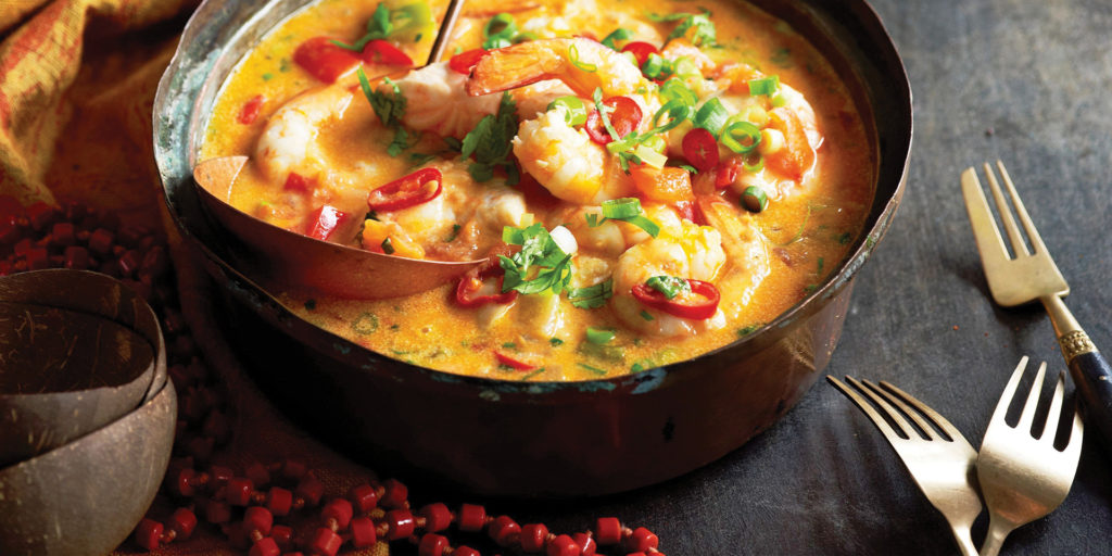 A rustic copper pot filled with vibrant orange-yellow Brazilian Moqueca, a seafood stew with plump shrimp, diced red peppers, and green herbs, served with a large serving spoon and two forks on a dark surface.