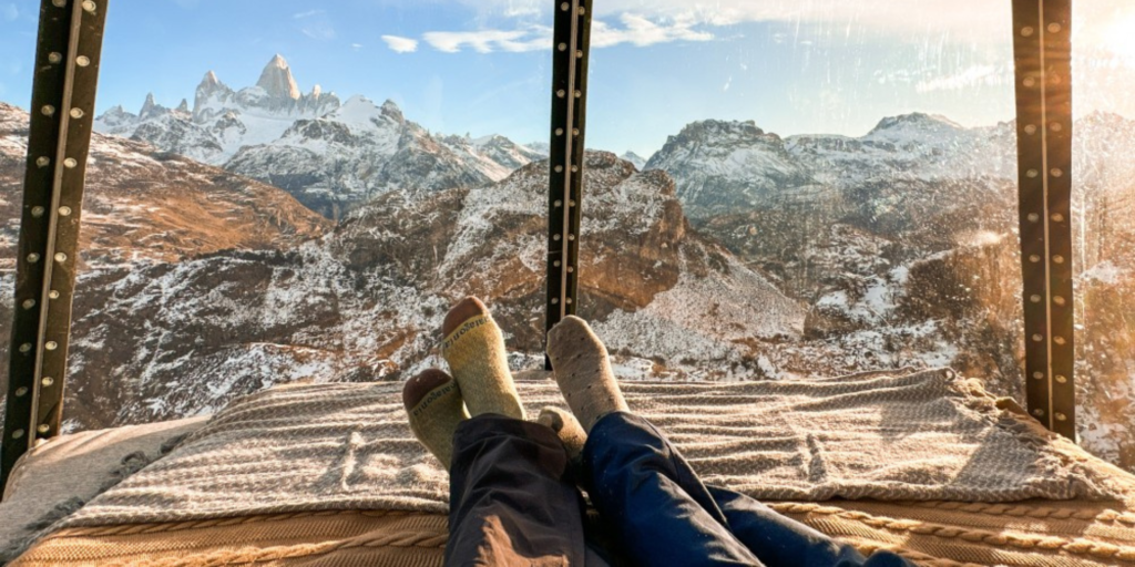 Two pairs of feet wearing socks resting on a striped blanket-covered bed, facing a large window revealing a panoramic view of snow-capped mountains and rocky terrain. The window frame is visible, suggesting an enclosed space.