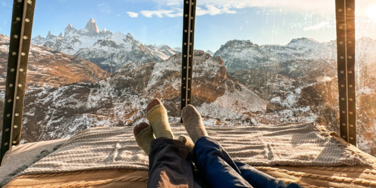 Two pairs of feet wearing socks resting on a striped blanket-covered bed, facing a large window revealing a panoramic view of snow-capped mountains and rocky terrain. The window frame is visible, suggesting an enclosed space.