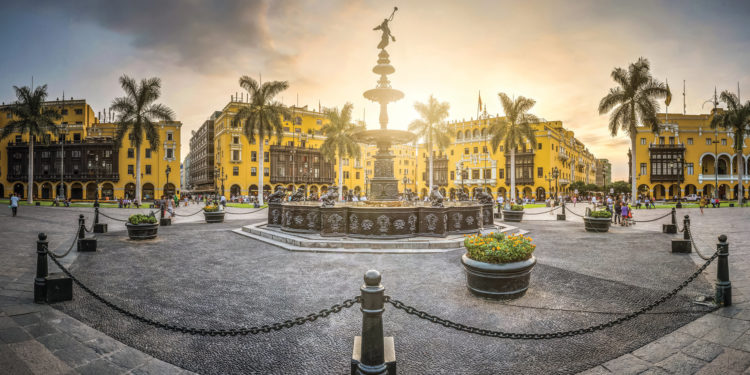 A panoramic view of Plaza de Armas in Lima, Peru, at sunset. A fountain stands at the center, surrounded by colonial buildings and palm trees.