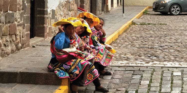 Three people in vibrant traditional clothing sit along a stone street in Cusco.
