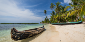 A wooden canoe on a white sandy beach with palm trees and turquoise water in the background.
