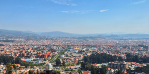 An aerial view of Cuenca, Ecuador, showing a sprawling cityscape with red-tiled roofs, surrounded by mountains under a clear blue sky.