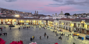 Quito plaza at twilight with people, buildings, and a church dome.