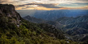 A panoramic view of Copper Canyon at sunset, with a cliff face in the foreground and layered mountains fading into the distance.