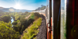 View from a train window of a long passenger train curving through a lush green valley with a river, mountains in the distance, and sunlight shining brightly.