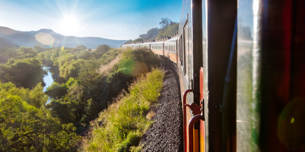 View from a train window of a long passenger train curving through a lush green valley with a river, mountains in the distance, and sunlight shining brightly.