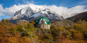 A medium shot of two green dome-shaped structures nestled amidst colorful autumn foliage. The domes feature wooden bases and triangular window panels. A snow-capped mountain is visible in the background, with a bright blue sky and fluffy white clouds.