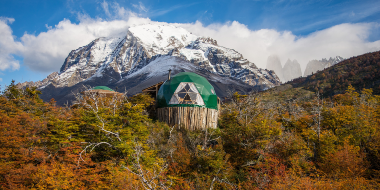 A medium shot of two green dome-shaped structures nestled amidst colorful autumn foliage. The domes feature wooden bases and triangular window panels. A snow-capped mountain is visible in the background, with a bright blue sky and fluffy white clouds.