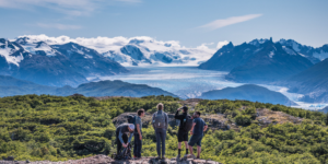 A group of people standing together on a grassy field in Patagonia, looking out at a vast landscape. In the background, the majestic Grey Glacier is visible with its icy blue surface and towering cliffs.