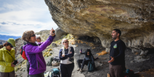 A group of people standing in front of a cave entrance, listening to a guide. The cave has rock art visible. The landscape is mountainous with vegetation.