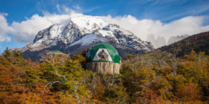 A medium shot of two green dome-shaped structures nestled amidst colorful autumn foliage. The domes feature wooden bases and triangular window panels. A snow-capped mountain is visible in the background, with a bright blue sky and fluffy white clouds.