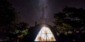A night shot of a dark dome with a brightly lit triangular window, surrounded by trees. The night sky above is filled with stars, including the Milky Way.