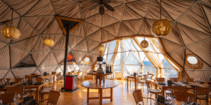 An interior shot of a geodesic dome used as a dining room. The dome has triangular glass panels, revealing a view of trees and another white dome outside. Inside, there is a set dining table with chairs, a couch, and wicker armchairs. The floor is made of polished wood.