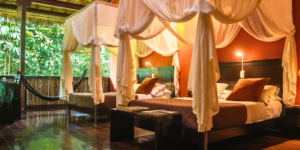 A room with two beds with draped canopies and a balcony overlooking a forest.