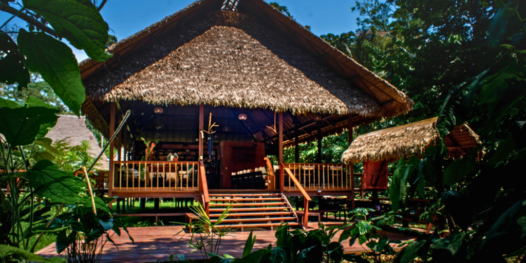 Exterior view of the Tambopata Research Center lobby, featuring a thatched roof, wooden construction, and tropical plants.