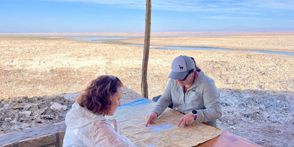 A traveller and a guide sit at a table with a large map spread out between them, while the guide points at specific locations on the map. They are in the vast salt plains of Atacama, with a stunning backdrop of salt flats and distant mountains under a clear blue sky.