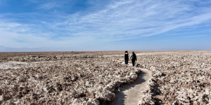 Two people walking along a narrow path through the rugged salt flats of Atacama under a clear blue sky. The vast, barren landscape of salt and rock extends to the horizon.