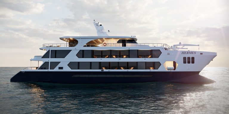 The image showcases the Hermes Mega Catamaran, a sleek and modern vessel with a predominantly white hull and a navy blue base. It's positioned in open water, likely at sunset, with a warm glow illuminating the sky. The catamaran's design emphasises spaciousness and comfort, with multiple decks and large windows.