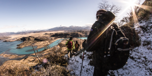 Hikers descend a snowy trail with a panoramic view of a turquoise river and mountains in Patagonia.