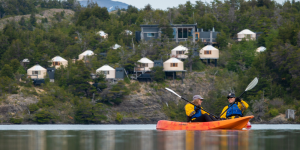 Two people kayaking on a calm lakes surrounded by lush greenery and mountains. Patagonia Camp yurts are perched on a hill behind them.