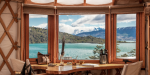 A cosy yurt interior with a large window offering a stunning view of a lake and mountains in Patagonia.