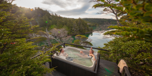 Two people relaxing in a hot tub on a wooden deck overlooking a beautiful lake surrounded by lush green forest and mountains.