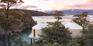 Two white yurts perched on stilts over a lake in Patagonia. The sky is pink and orange at sunset.