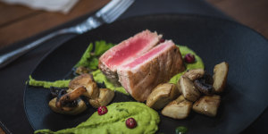 A plate of seared tuna steak served with sautéed mushrooms, a green pea purée, and a red peppercorn sauce.