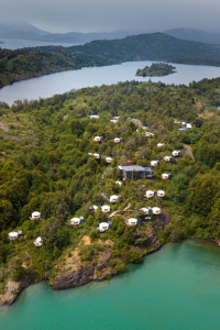 An aerial view of Patagonia Camp, a luxury eco-lodge nestled on the shores of a turquoise lake surrounded by lush green forests and mountains.