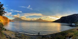 Two people walking on a beach with a lake and mountains in the background.