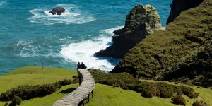 Two people sitting on a boardwalk overlooking a dramatic coastline.