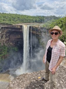 Heather standing on a rocky outcrop overlooking a massive waterfall cascading into a gorge, with lush rainforest in the background.