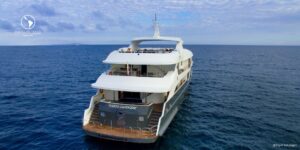 A luxury yacht, the Infinity, anchored in blue waters in the Galapagos. The yacht has a dark grey hull, a white upper deck with windows, and a crane on the side.